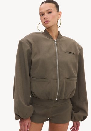 Femme avec de grandes boucles d'oreilles en or porte une veste zippée oversize vert olive et un short assorti sur un fond clair uni.
