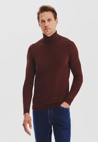 Brązowy sweter z golfem wykonany z gładkiego, dopasowanego materiału, z długimi rękawami i przylegającym fasonem, w połączeniu z ciemnoniebieskimi dżinsami.