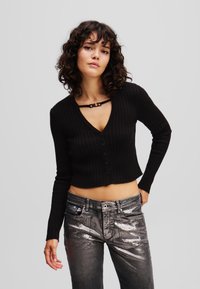 Černý žebrovaný pletený cardigan s výstřihem do V a zapínáním na knoflíky, kombinovaný se stříbrnými metalickými džíny. Má jedinečný detail pod výstřihem.