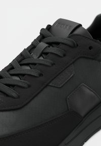 Baskets noires en matériau synthétique texturé, avec lacets plats et design épuré. Logo en relief notable sur le côté et accents subtils.