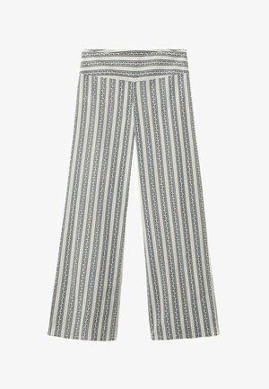 Mladá žena nosí čierno-biely pruhovaný halter top a široké nohavice, stojac na jednoduchom bielom pozadí.