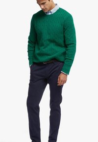 Grüner kabelgestrickter Pullover mit rundem Ausschnitt, kombiniert mit marineblauen Hosen. Der Pullover weist strukturierte Muster und gerippte Bündchen auf.