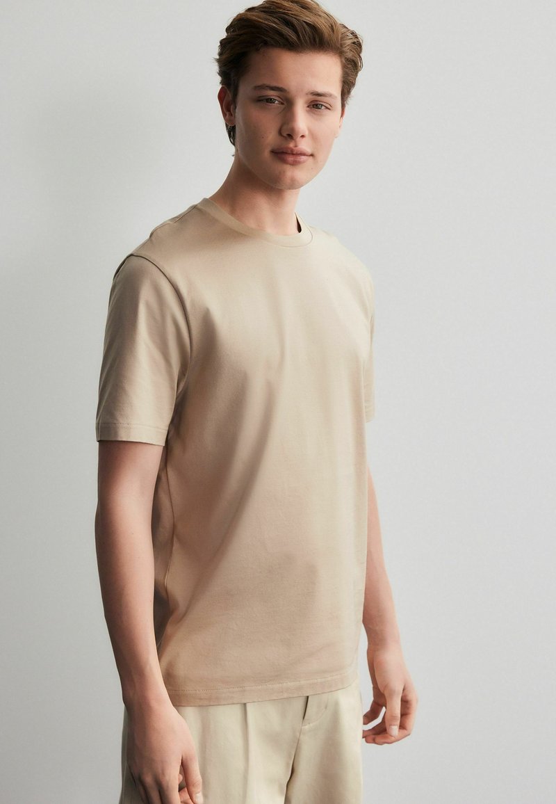 Beige bomuld t-shirt med klassisk rund hals, korte ærmer og lige skæring. Glat tekstur, minimalistisk design, ingen synlige mønstre.