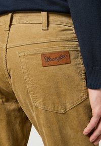 Pantaloni in velluto a coste marroni con una patch in pelle 'Wrangler' sulla tasca posteriore, caratterizzati da una texture a coste e dettagli cuciti.