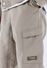 Pantalones cargo beige con un bolsillo lateral que lleva una etiqueta rectangular marcada con "PROJECTX PARIS". Tejido suave y duradero con textura mínima.
