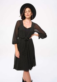 Zwarte jurk met stippen, een strik taille, doorzichtige lange mouwen en een uitlopende rok. Het patroon bevat gouden stippen en heeft een gelaagd effect.