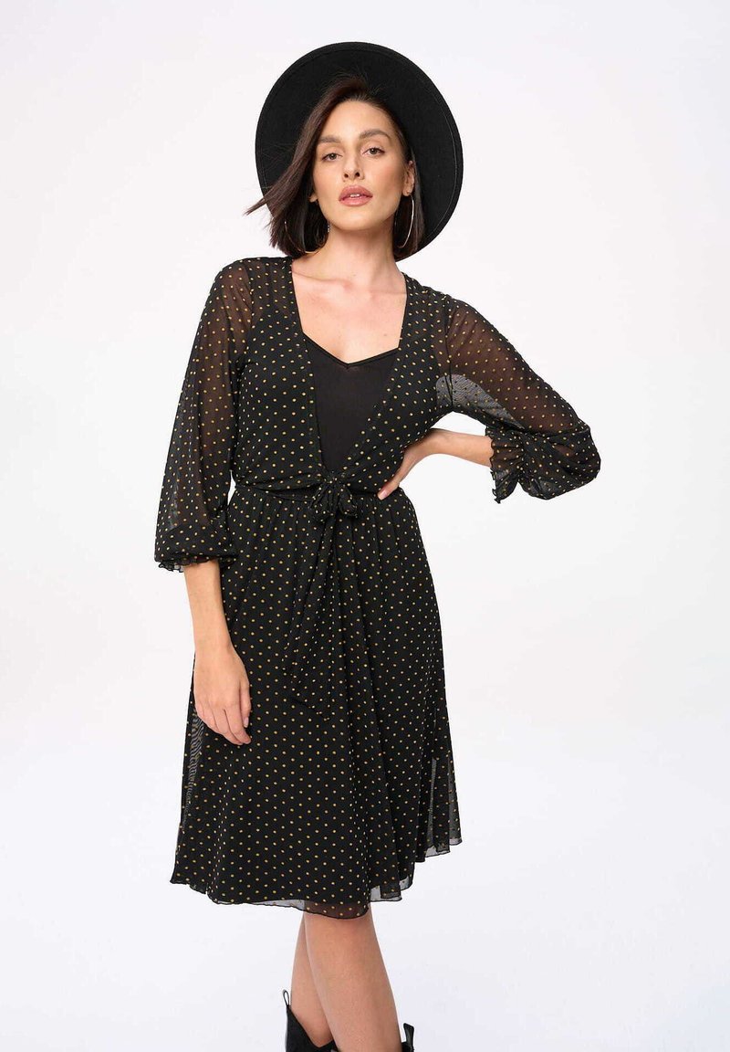 Zwarte jurk met stippen, een strik taille, doorzichtige lange mouwen en een uitlopende rok. Het patroon bevat gouden stippen en heeft een gelaagd effect.