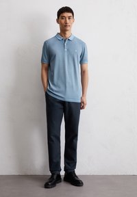 Lichtblauwe polo gemaakt van stof met een gladde textuur, gecombineerd met donkere broeken en zwarte veterschoenen. Eenvoudig logo-detail op het shirt.