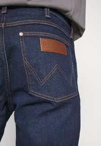 Temno modri kavbojki iz denima z usnjeno oznako "Wrangler." Imajo zlat šiv in značilen dizajn zadnjega žepa.