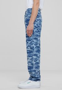 Light blue camouflage pants made of denim, featuring a straight fit, five-pocket design, and subtle stitching details, paired with white sneakers.