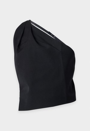 Femme portant une veste courte argentée à sequins sur un haut noir, un jean bleu foncé et des talons noirs ouverts, debout devant un fond uni.