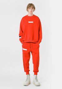 Sudadera roja con una etiqueta plateada, combinada con pantalones jogger rojos que presentan bolsillos laterales y un diseño en forma de llama. Llevado con botas blancas.