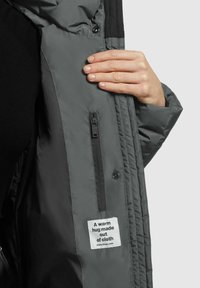 Grijze gewatteerde jasinterieur met een ritszak, zwarte stofaccenten en een label met de tekst: "Een warme omhelzing gemaakt van stof."