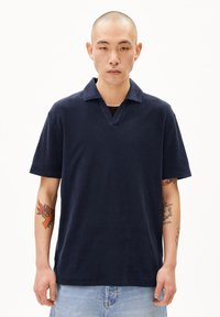Marineblauw polo shirt met korte mouwen, een gestructureerde afwerking en een V-hals. Model toont zichtbare tatoeages op de arm.