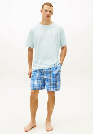 Junger Mann steht barfuß, trägt ein hellblaues T-Shirt mit kurzen Ärmeln und blau karierte Shorts vor einem schlichten weißen Hintergrund.