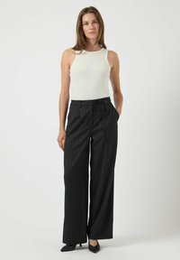 Vrouw die staat en een witte mouwloze top, zwarte high-waisted wijde gestreepte broek en zwarte puntige schoenen draagt.