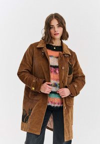 Brązowa kurtka z kordurowej tkaniny z guzikami, przednimi kieszeniami i grafiką lisa po boku, noszona na multikolorowym swetrze dzianinowym.