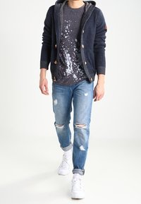Marinblå stickad cardigan med träknappar över en grå t-shirt med splatter-mönster, i kombination med ljusa, trasiga denimjeans och vita sneakers.