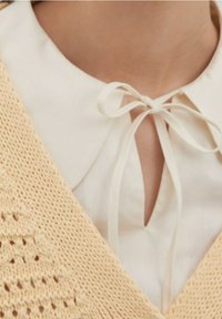 Weiße Bluse mit gebundenem Schleifenkragen unter einem beige gestrickten Pullover, der den oberen Brust- und Halsbereich zeigt.