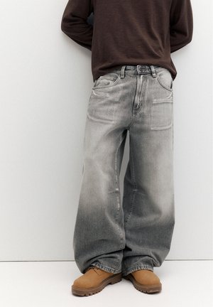 Személy kopott, széles szárú szürke farmert, barna hosszú ujjú felsőt és világosbarna munkavédelmi bakancsot visel, sima világos háttér előtt áll.