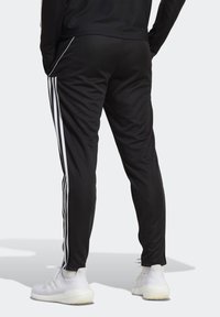 Svarta träningsbyxor med vita sidostrecker, smal passform, elastisk midja och dragkedjeslut, kombinerat med vita sneakers.