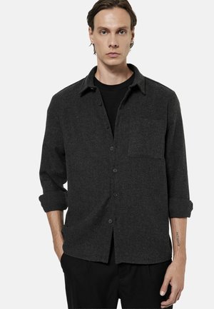 Młody mężczyzna w ciemnoszarej koszuli z guzikami i podwiniętymi rękawami oraz czarnych spodniach, stojący na jednolitym tle.