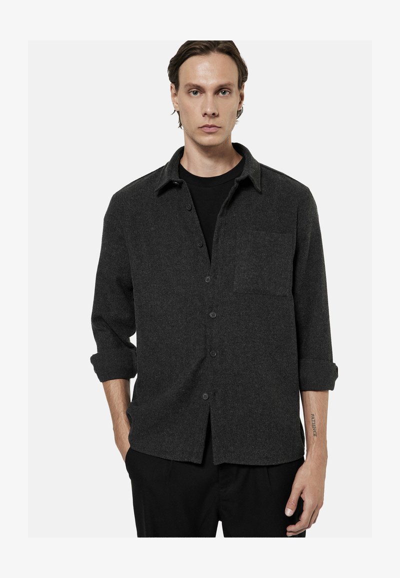 Jeune homme portant une chemise gris foncé à boutons avec les manches retroussées et un pantalon noir, debout devant un fond uni.