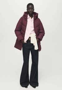 Vínová prošívaná bunda s kapucí, pod ní světle růžová košile, tmavé rozšířené džíny a bílý svetr uvázaný v pase. Celý vzhled doplňují černé podpatky.