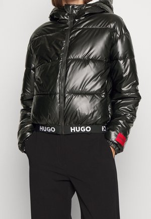 Doudoune noire brillante avec capuche, fermeture éclair frontale, ourlet élastique marqué HUGO et patch rouge sur la manche, portée avec un pantalon noir.