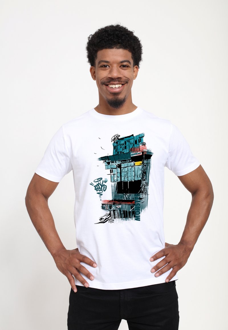 Hombre sonriente con cabello corto y rizado, vistiendo una camiseta blanca con un diseño de edificio estilo graffiti en azul y negro, que incluye texto y pájaros.