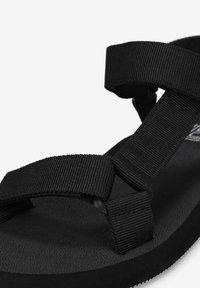 Sandalia negra con suela de caucho texturizada y amplias tiras de nailon trenzado con acentos de botón, diseñada para comodidad y durabilidad.