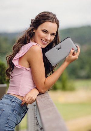 Gray textured wallet with contrasting black zipper and embossed logo. Model with long hair holds wallet against a natural background.