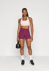 Črna kettlebell, ki jo drži v stoječem položaju. Ima na sebi svetlo rožnato športno modrček in temno vijolične kratke hlače z visokim pasom, v kombinaciji s črnimi supergami in belimi nogavicami.