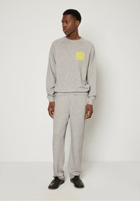 Siv sveder iz bombaža in ujemajoče se športne hlače imata spredaj rumeni logotip. Model nosi črne čevlje, ima sproščen kroj in rebraste manšete.