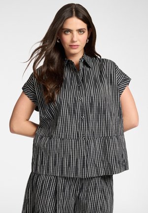 Kobieta o długich brązowych włosach, ubrana w czarno-białą koszulę w pionowe paski z krótkim rękawem zapinaną na guziki oraz dopasowane spodnie, stojąca z rękami za plecami.