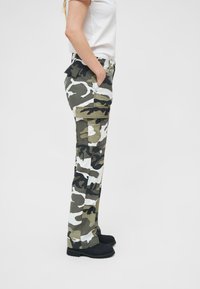 Vihreät, mustat ja valkoiset camoleggings-housut. Sisältää useita taskuja, leveät lahkeet ja tekstuuripinta, jossa on voimakas camouflage-kuvio.