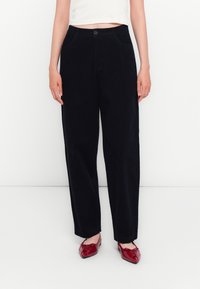 Femme portant un pantalon noir taille haute à jambes droites, un top court blanc et des ballerines rouges en cuir verni, debout sur fond blanc.