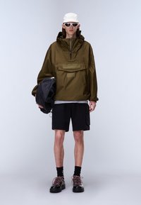 Person iført olivengrøn hættetrøje, sorte shorts, hvid kasket, hvide solbriller, sorte sokker, sorte sko, der holder en sort rygsæk.