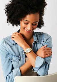 Orologio da polso dorato con quadrante rotondo, indicatori delle ore verdi e bracciale metallico. Indossato su un polso con uno sfondo di camicia in denim.