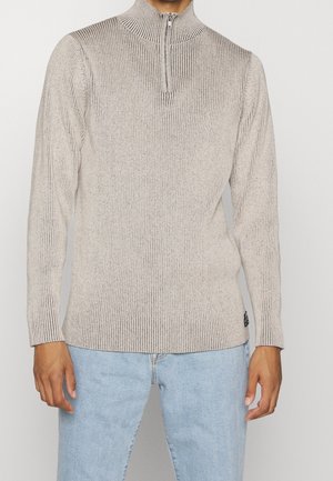Maglione beige lavorato a maglia con colletto alto e chiusura a zip a un quarto. Presenta una consistenza a coste e maniche lunghe, abbinato a jeans azzurri chiari.