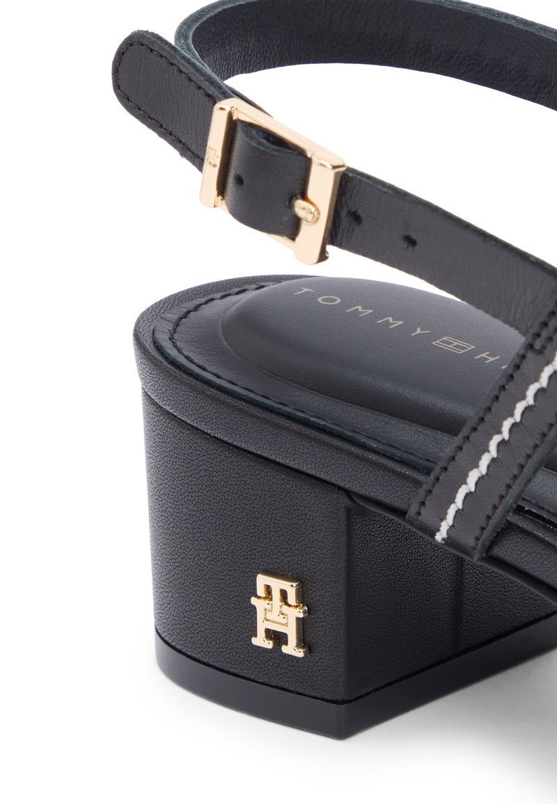 Sandalia de tacón de cuero negro con correa de hebilla dorada, costuras blancas y logo dorado de Tommy Hilfiger en el talón.