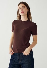 Rjava kratka majica s kratkimi rokavi in okroglim izrezom, gladke teksture in prilegajočega kroja, v kombinaciji s temnimi kavbojkami.