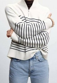 Maglione bianco a costine con strisce nere, collo zip e vestibilità ampia. Abbinato a jeans di denim azzurri. Tessuto testurizzato con motivi a contrasto.