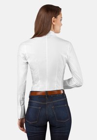 Weißes, tailliertes Hemd mit langen Ärmeln, glatter Textur und subtiler Schattierung, kombiniert mit dunkelblauen Jeans und einem braunen Ledergürtel.