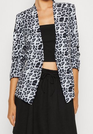 Nainen pukeutunut harmaaseen ja mustaan leopardikuvioiseen bleiseriin mustan putkikorin ja mustien naruhousujen päällä valkoista taustaa vasten.