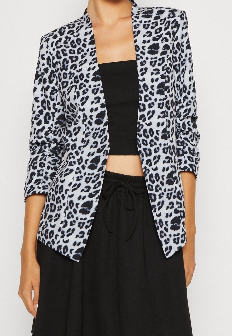 Femme portant un blazer imprimé léopard gris et noir sur un top bandeau noir et un pantalon noir à cordon, sur fond blanc.