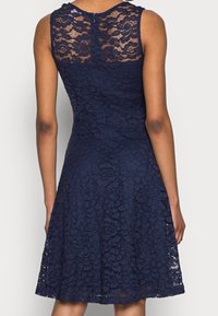 Mörkblå spets ärmlös klänning med blommönster, figursydd midja och utsvängd kjol, visad bakifrån på en stående person.