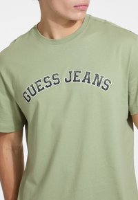 Olive spalvos medvilnės marškinėliai su kreiva tamsia "GUESS JEANS" grafika, apvalia iškirpte ir trumpomis rankovėmis. Lygus paviršius.