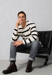 Pasiasta koszula z długim rękawem w kolorze kremowym i czarnym, szare dżinsy, oraz brązowe skórzane buty na sznurówki, siedząca na czarnym krześle przy fakturowanej ścianie.