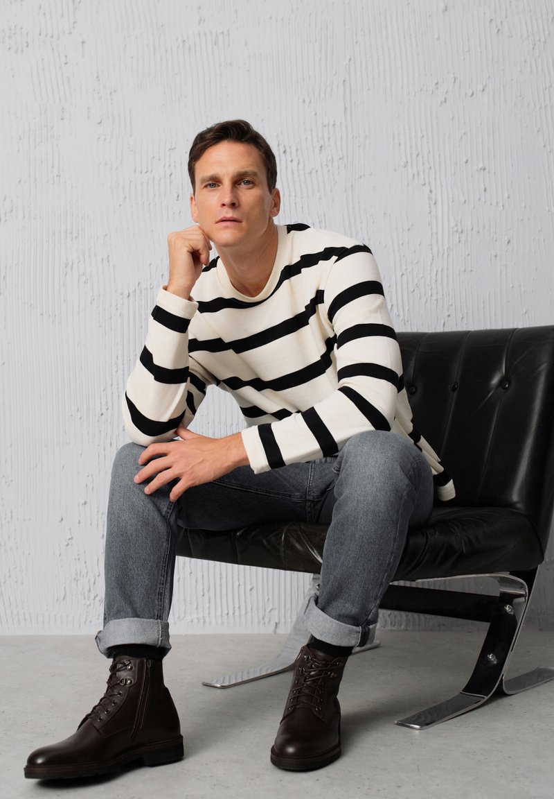 Pasiasta koszula z długim rękawem w kolorze kremowym i czarnym, szare dżinsy, oraz brązowe skórzane buty na sznurówki, siedząca na czarnym krześle przy fakturowanej ścianie.